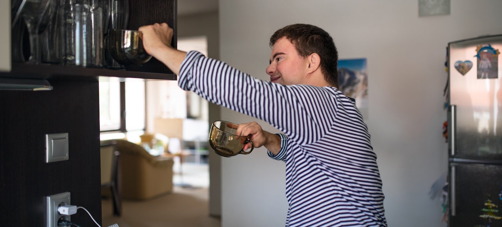 Ein junger Mann mit Down-Syndrom räumt in einer hellen Küche dunkle Glastassen in einen Oberschrank ein.