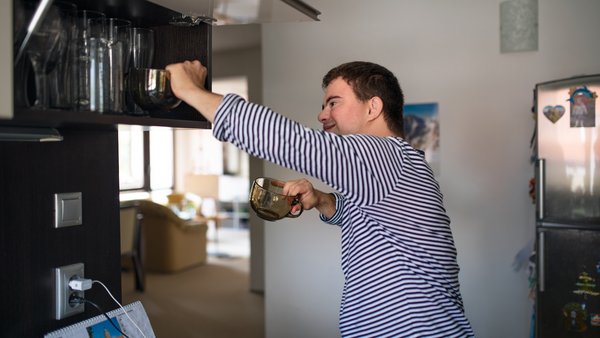 Ein junger Mann mit Down-Syndrom räumt in einer hellen Küche dunkle Glastassen in einen Oberschrank ein.