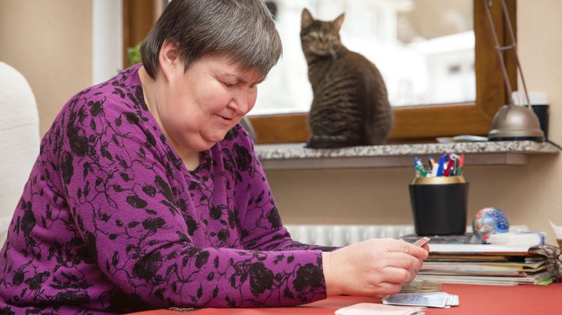 Frau mit Behinderung sitzt am Tisch und spielt Karten. Eine Katze sitzt auf der Fensterbank.