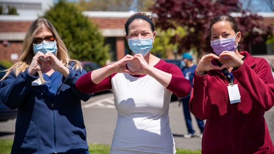 Drei junge Frauen mit Maske zeigen mit ihren Händen ein Herz.
