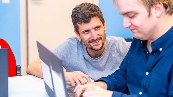 Ein junger Mann sitzt an einem Laptop und tippt, während ein zweiter Mann mit Bart im grauen T-Shirt lächelnd von der Seite zuschaut und ihn unterstützt.