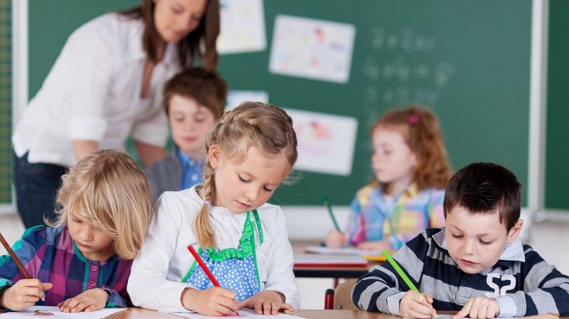 Junge Schulkinder lösen Aufgaben im Heft. Die Lehrerin erklärt einem Kind etwas.
