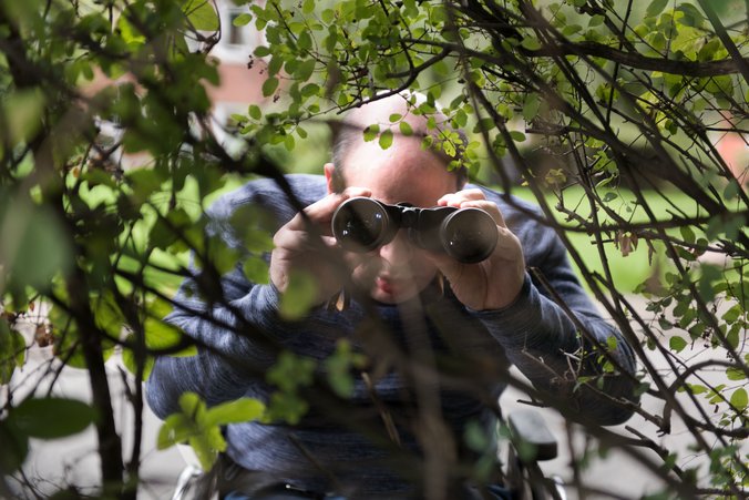 Ein Mann richtet ein Fernglas auf etwas, das er zwischen den Zweigen einer Hecke sieht.