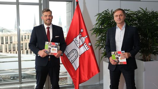 Falko Droßmann, Bezirksamtsleiter Hamburg-Mitte, und Dr. Andreas Theurich, Vorsteher des Rauhen Hauses, stehen neben einer Hamburg-Fahneund stellen das neue Buch "Lebenswelten im Dialog" vor.