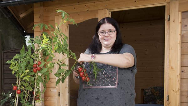 Eine Frau mit dunklen langen Haaren steht vor einer Holzhütte und schneidet einen Tomatenstrauch.