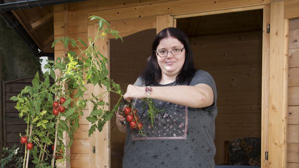 Eine Frau mit dunklen langen Haaren steht vor einer Holzhütte und schneidet einen Tomatenstrauch.