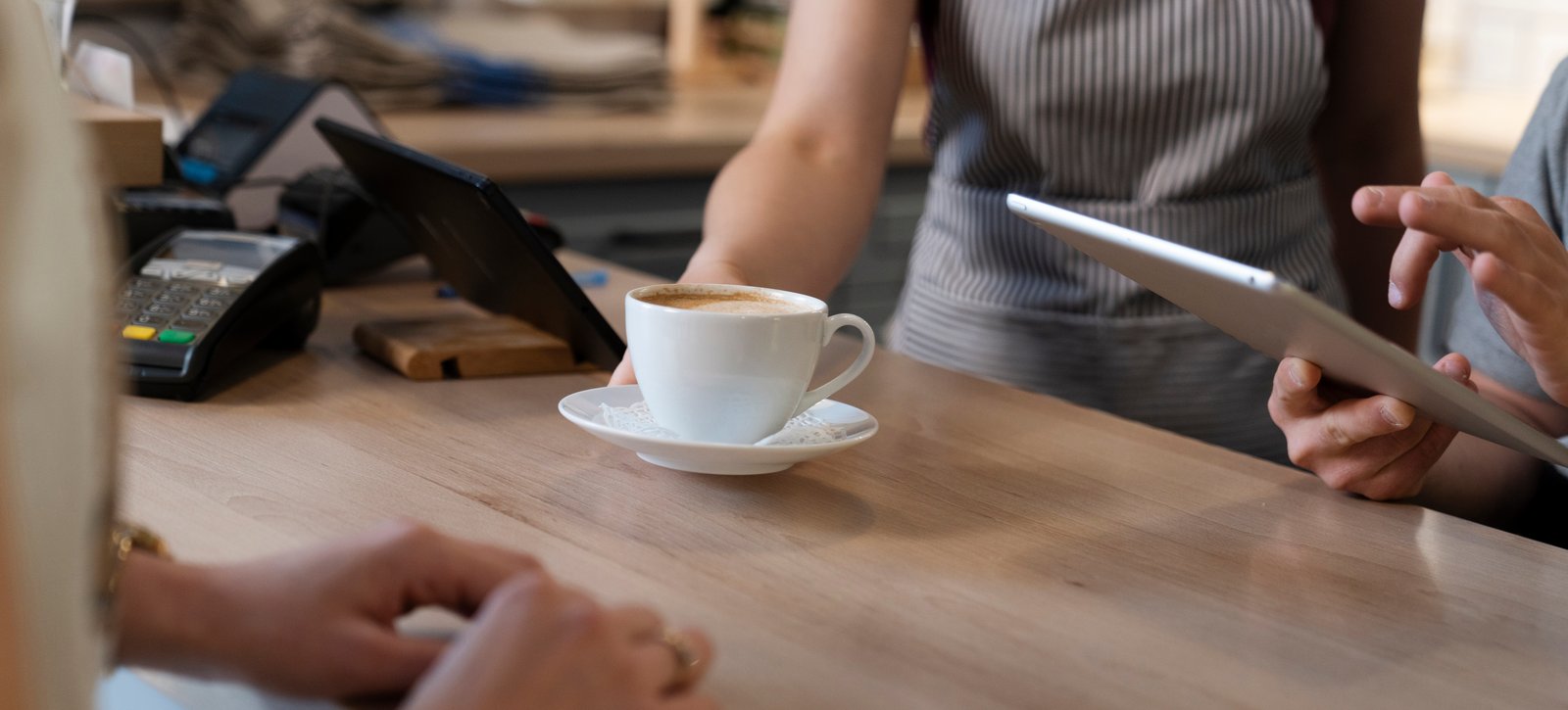 Das Bild zeigt eine klassische Szene in einem Café, in der ein Barista einen Kaffee serviert, während ein Kunde oder eine andere Person ein Tablet bedient. Die Kaffeetasse auf dem Tresen ist ein typisches Modell für die Gastronomie.