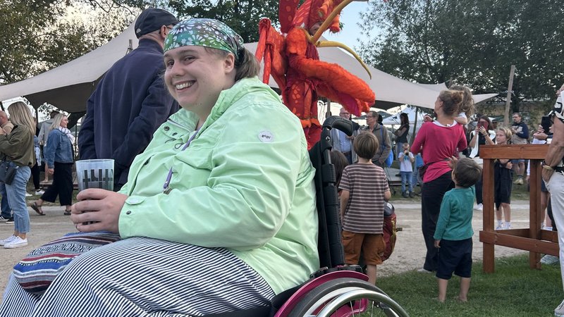 Eine Frau sitzt im Rollstuhl und lacht. Im Hintergrund sieht man das Festival.