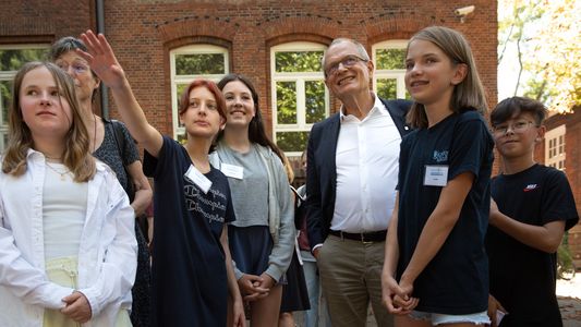 Eine Gruppe von Schulkindern steht mit Ulrich Lilie, Diakonie-Präsident, vor dem Paulinum auf dem Stiftungsgelände und erklären ihm etwas.